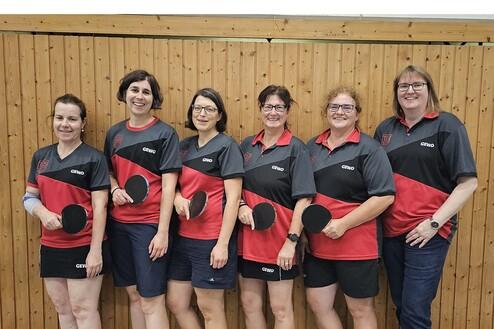 Sechs Frauen in Tischtenniskleidung posieren lächelnd mit Schlägern vor einer Holzwand.