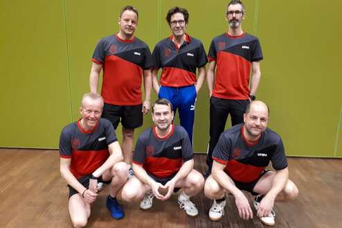 Sechs Männer in roten und grauen Sporttrikots stehen vor einer grünen Wand in einer Sporthalle.