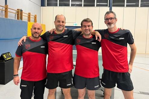 Vier Männer in roten Tischtennis-Trikots stehen nebeneinander, lächeln und umarmen sich in einer Sporthalle.