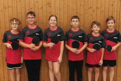 Sechs Kinder in roten Tischtennistrikots stehen vor einer Holzwand und halten Tischtennisschläger.