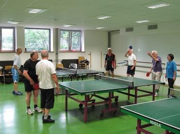 Gruppe älterer Menschen spielt Tischtennis in einer Halle mit mehreren Tischtennisplatten und großen Fenstern im Hintergrund.