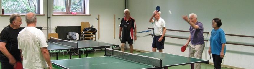Gruppe &auml;lterer Menschen spielt Tischtennis in einer Halle mit mehreren Tischtennisplatten und gro&szlig;en Fenstern im Hintergrund.
