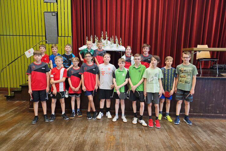 Gruppe von 16 Tischtennisspielern in Sportkleidung vor einer Bühne mit Pokalen, in einem großen Saal.