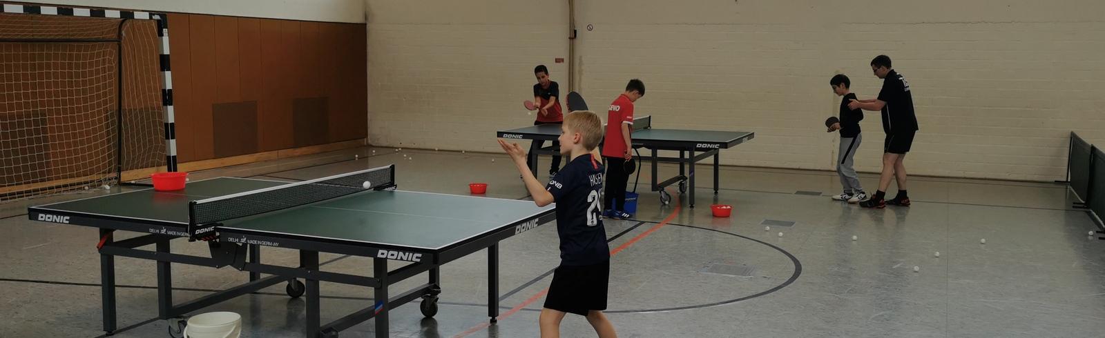 Kinder trainieren Tischtennis in einer Sporthalle mit zwei Tischtennisplatten und einem Basketballkorb im Hintergrund.