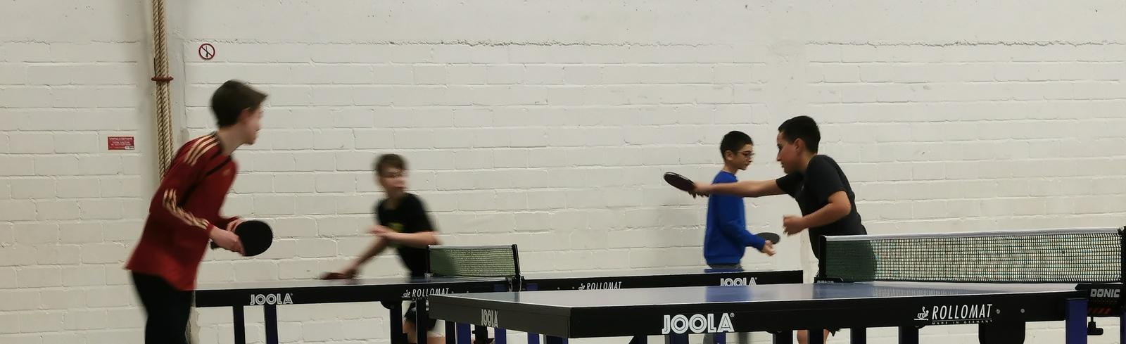 Vier Kinder spielen Tischtennis in einer Halle mit zwei Tischtennisplatten und einem Ball auf dem Boden.