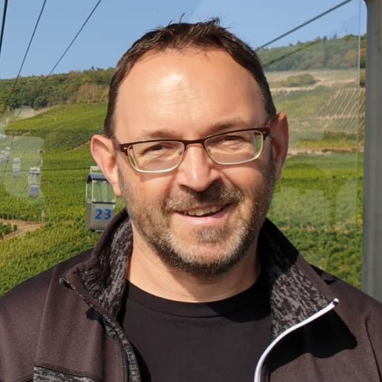 Mann mit Brille lächelt vor einer Grünen Weinberglandschaft, während er in einer Seilbahn sitzt.