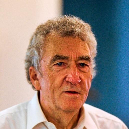 Porträt eines älteren Mannes mit grauen Haaren und einem weißen Hemd, der in einem hellen Raum spricht.