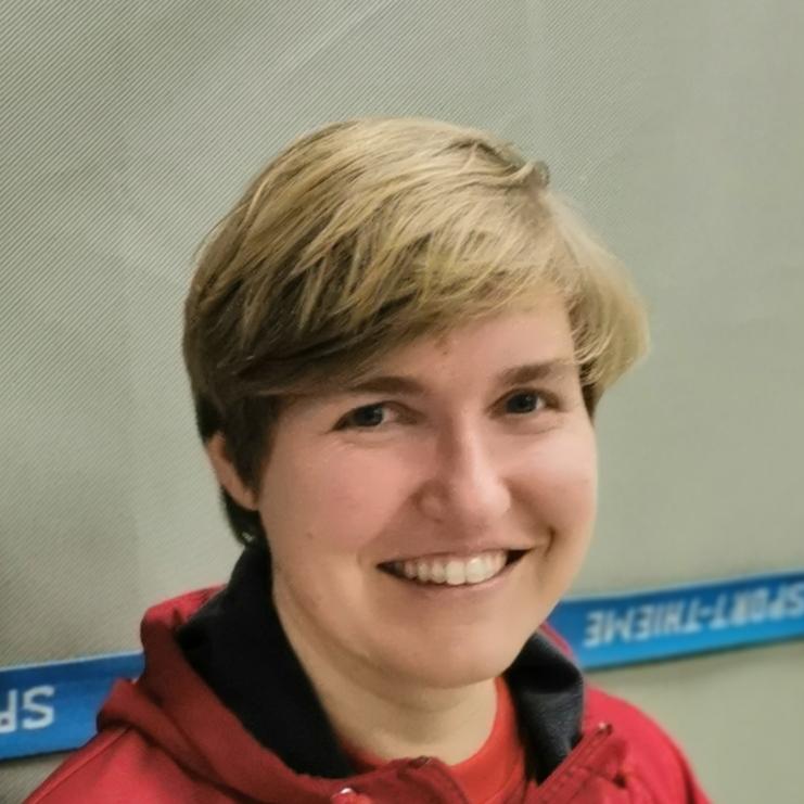 Lächelnde Person mit kurzem, blondem Haar, trägt eine rote Jacke und steht vor einer grauen Wand.