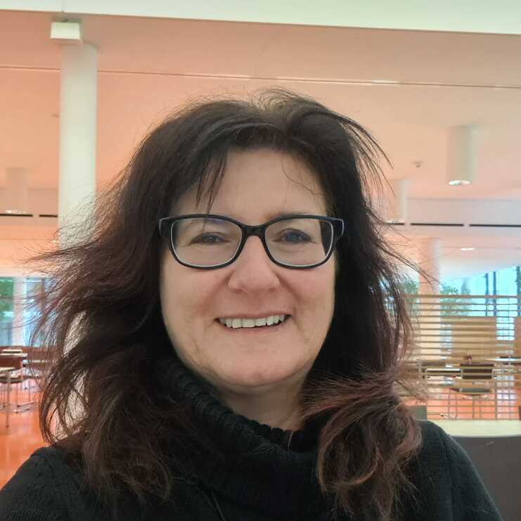 Portrait einer lächelnden Frau mit dunklem Pullover in einem modernen, hellen Innenraum mit Möbeln im Hintergrund.