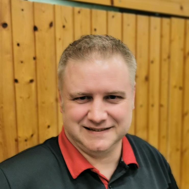 Lächelnder Mann in einem schwarzen und roten Sportpoloshirt mit dem Logo von TuS Griesheim vor einer Holzwand.