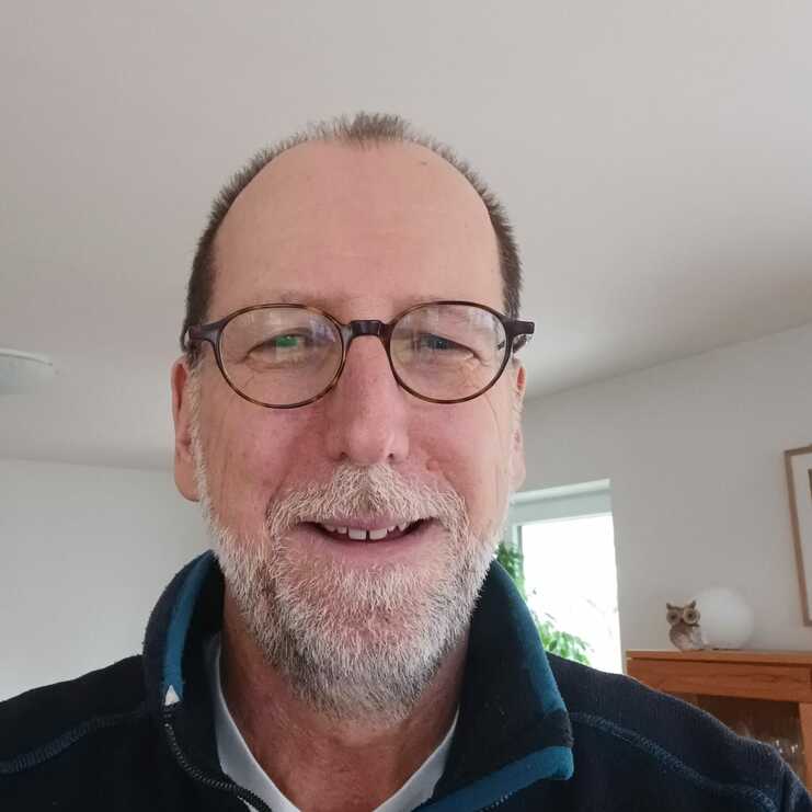 Selbstporträt eines Mannes mit Brille, kurzer grauer Bart und dunkler Jacke, in einem hellen Raum mit Pflanzen im Hintergrund.