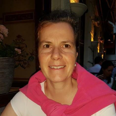 Frau mit kurzen Haaren und rosa Pullover, sitzend in einem Restaurant mit dekorativer Blumendeko im Hintergrund.