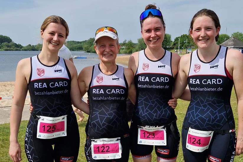 Vier Frauen in Triathlonanzügen stehen lächelnd am Ufer eines Gewässers, umgeben von grüner Landschaft.