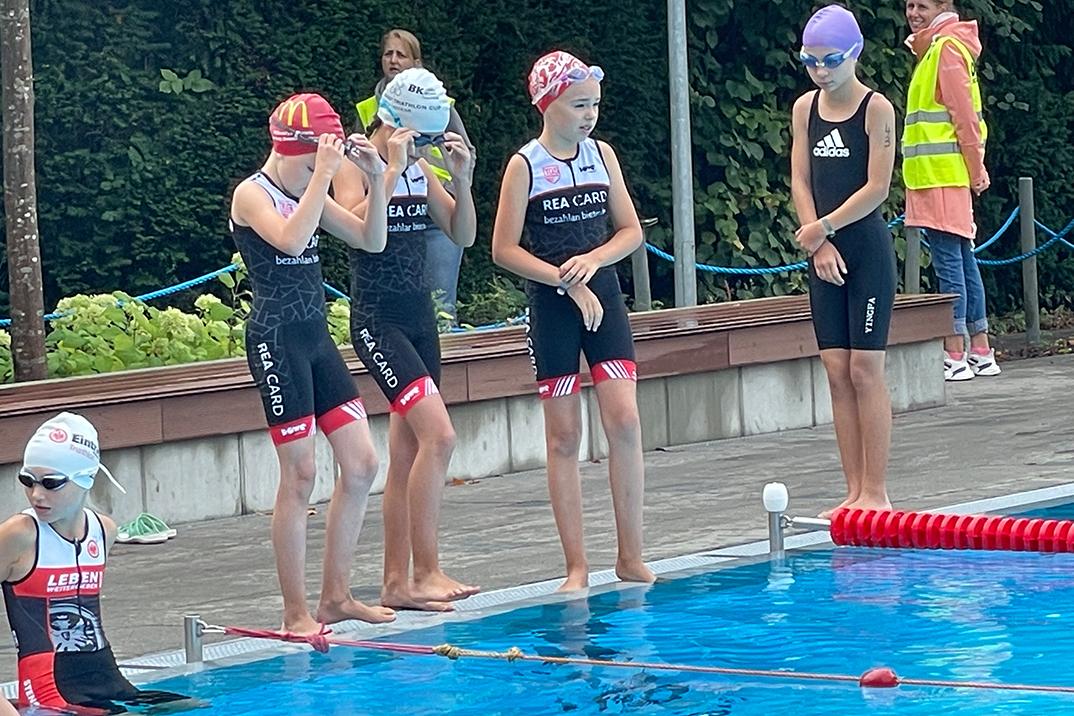Vier Schwimmerinnen in Badeanzügen und Badekappen stehen am Beckenrand und bereiten sich auf ihren Wettkampf im Schwimmbad vor.