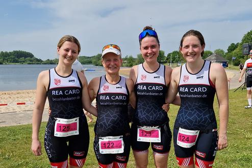 Vier Frauen in Triathlonanzügen stehen lächelnd am Ufer eines Gewässers, umgeben von grüner Landschaft.