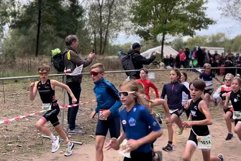 Eine Gruppe von Kindern läuft einen Triathlon-Wettbewerb, umgeben von Zuschauern und Absperrungen.