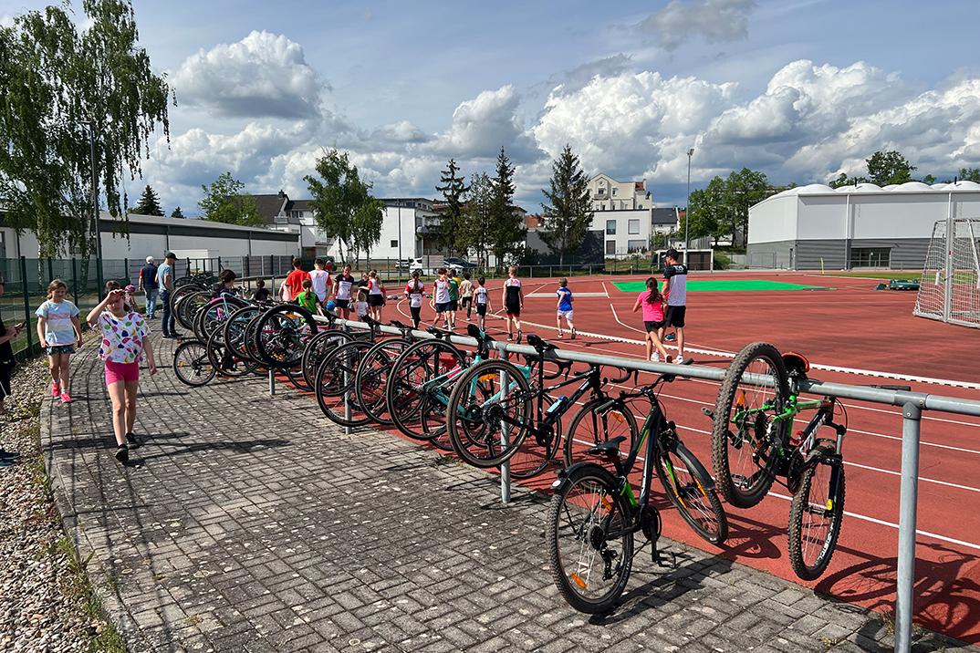 Junge Menschen und Fahrräder stehen an einer Sportanlage mit roten Laufbahnen und Wolken am Himmel.