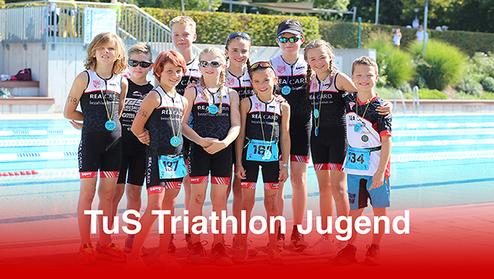 Gruppe von jungen Sportlern in Triathlonanzügen am Schwimmbecken, einige mit Medaillen um den Hals.
