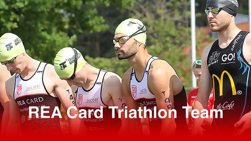 Fünf Männer in Schwimmkleidung und Badekappen stehen für einen Triathlon bereit, mit Blick auf die Strecke.