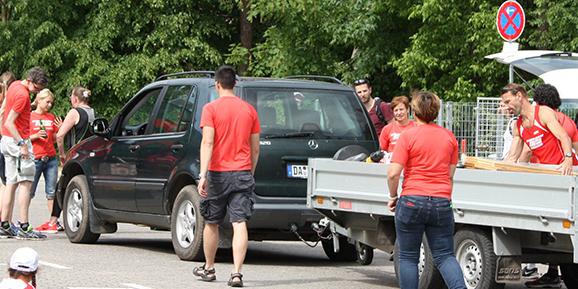 Mehrere Personen in roten T-Shirts stehen auf einem Parkplatz neben Autos und einem Anh&auml;nger, umgeben von B&auml;umen.