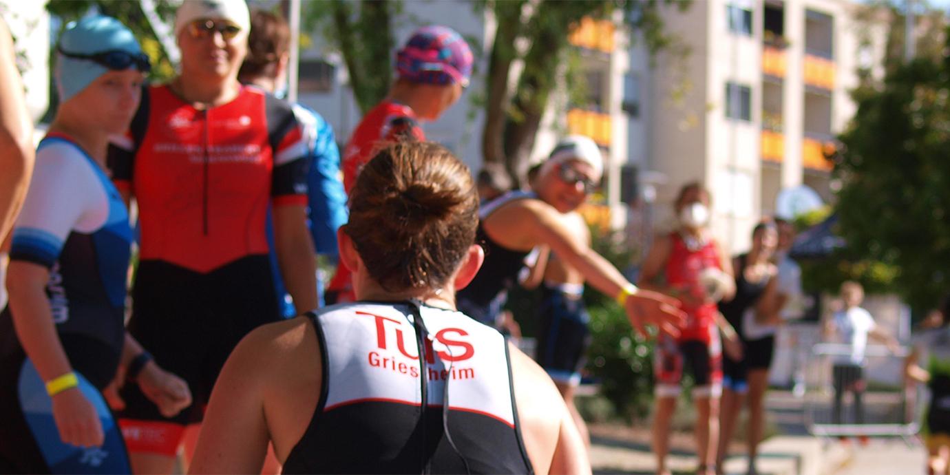 Gruppe von Triathleten in Sportkleidung vor einem Wettkampf, einige winken oder interagieren miteinander.