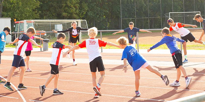 Gruppe von Kindern beim Dehnen auf einer Laufbahn, umgeben von grüner Natur und Sporteinrichtungen.