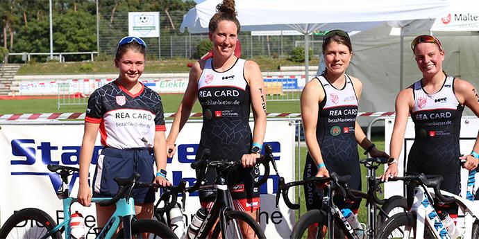 Vier Frauen stehen neben ihren Fahrr&auml;dern auf einem Sportgel&auml;nde, mit Werbebannern im Hintergrund.