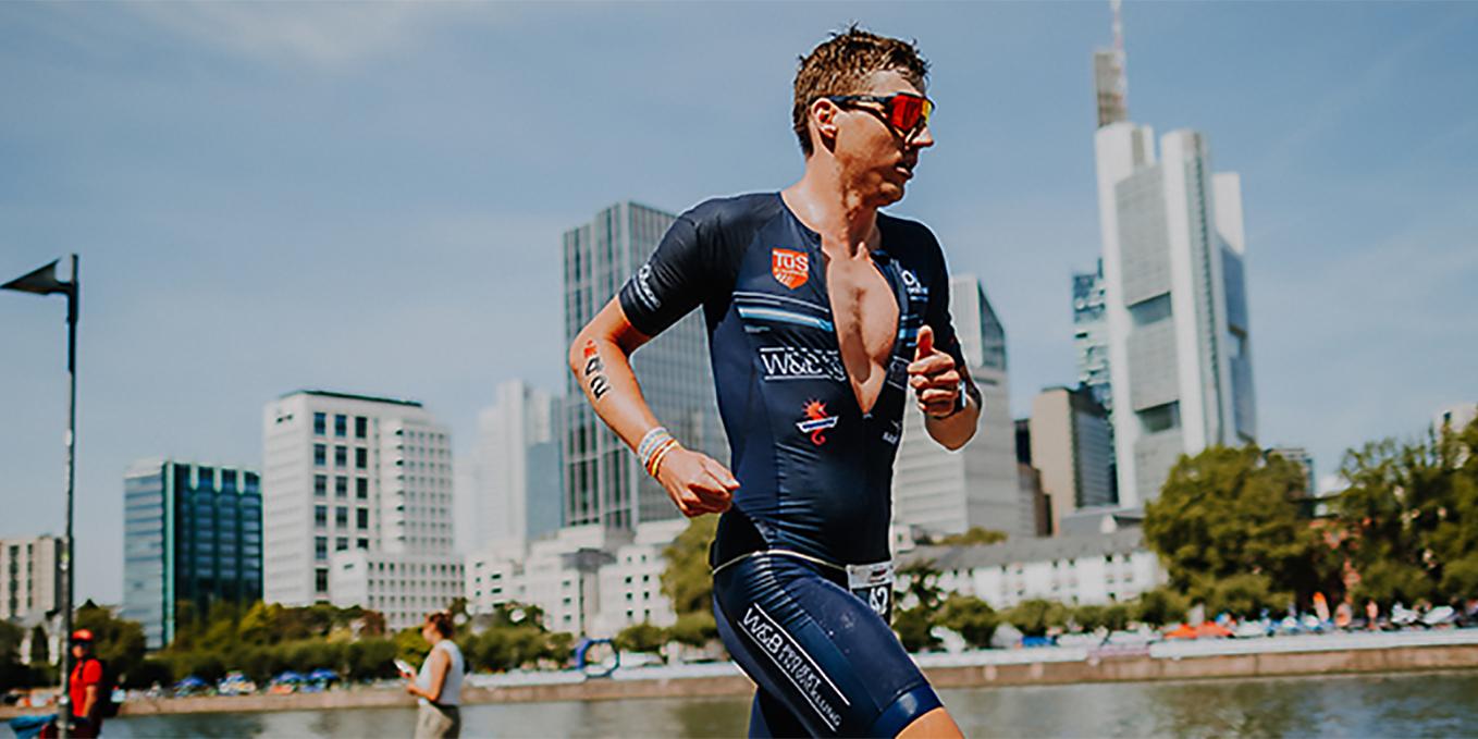 Ein Triathlet l&auml;uft am Wasser entlang, moderne Hochh&auml;user sind im Hintergrund sichtbar.