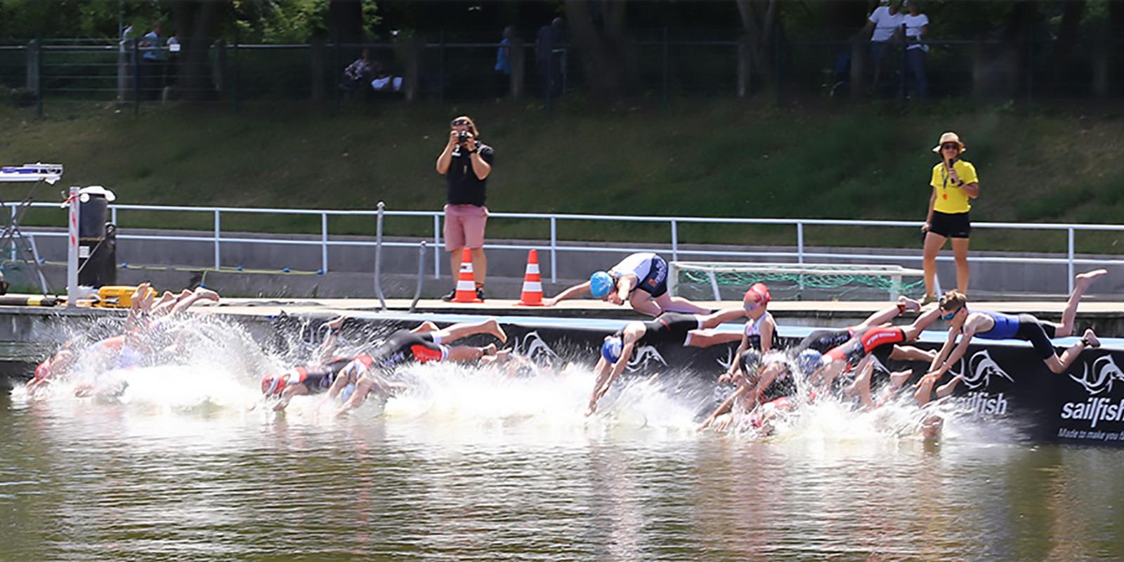 Athleten springen ins Wasser von einem Steg w&auml;hrend eines Wettkampfs, Zuschauer und Kameras im Hintergrund sichtbar.