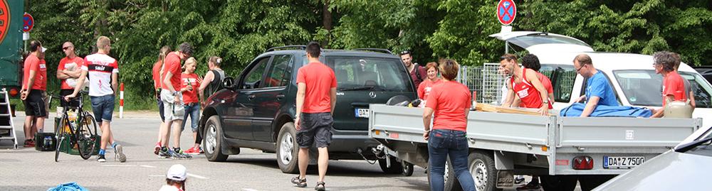 Mehrere Personen in roten T-Shirts stehen auf einem Parkplatz neben Autos und einem Anhänger, umgeben von Bäumen.