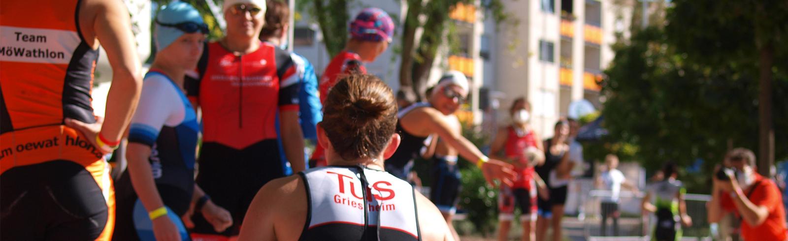 Gruppe von Triathleten in Sportkleidung vor einem Wettkampf, einige winken oder interagieren miteinander.