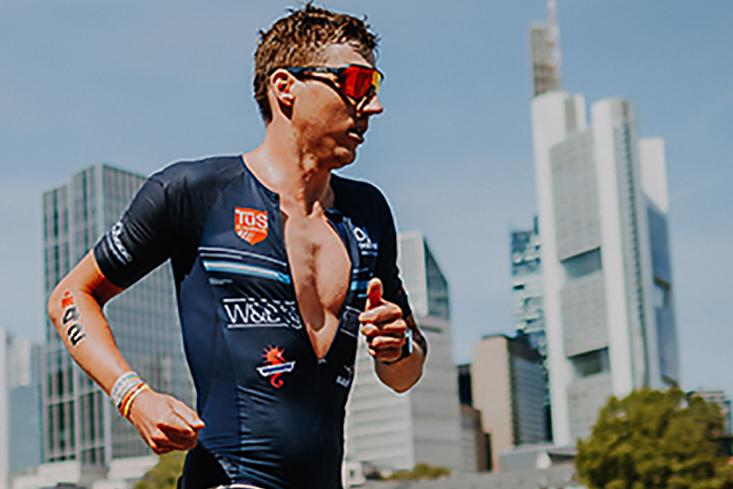 Ein Triathlet läuft am Wasser entlang, moderne Hochhäuser sind im Hintergrund sichtbar.