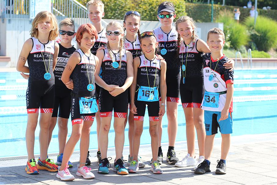 Gruppe von zehn Kindern in Sportanzügen, lächelnd, vor einem Schwimmbecken, einige mit Medaillen um den Hals.