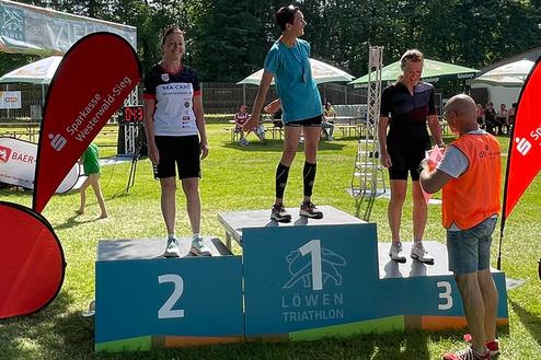 Drei Sportlerinnen stehen auf dem Siegertreppchen des Löwen Triathlons, während ein Veranstalter Preisunterlagen überreicht.