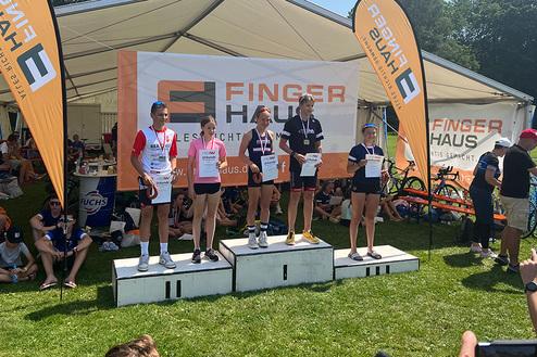 Siegerpodium mit fünf Athleten, Medaillen, Urkunden und Banner in einem festlichen Zelt bei sonnigem Wetter.