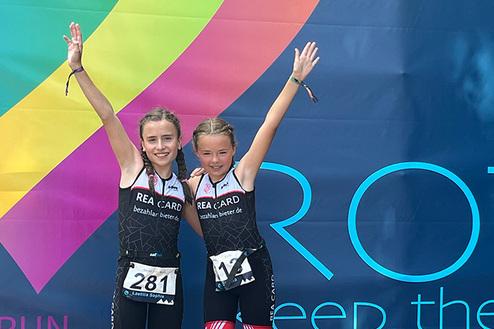Zwei fröhliche Mädchen in Sportanzügen posieren mit erhobenen Armen vor einer bunten Werbefläche.