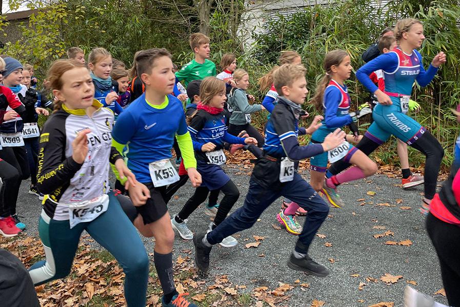 Gruppe von Kindern in Sportkleidung beim Laufen auf einem Weg, umgeben von Bäumen und herbstlichem Laub.