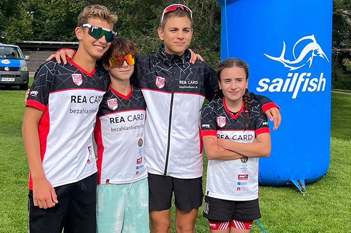 Vier Jugendliche in Sportkleidung posieren auf einer grünen Wiese vor einem blauen Inflatable Banner.