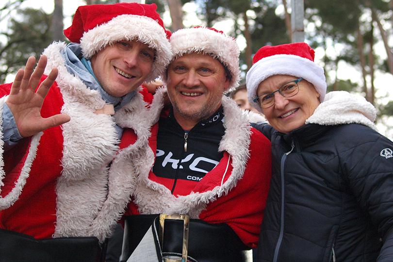 Drei Personen in Weihnachtsmann-Kostümen lächeln und posieren im Freien vor einem Stand und Bäumen.