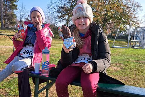 Zwei Mädchen sitzen auf einer Bank, essen Snacks und genießen den Sonnenschein auf einem Sportplatz.
