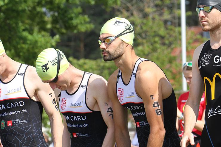 F&uuml;nf Triathleten in Neoprenanz&uuml;gen stehen startbereit, w&auml;hrend sie in die Kamera blicken.