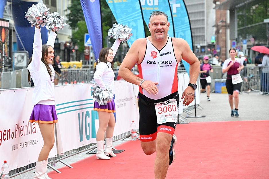 Mann im Wettkampfanzug sprintet auf roten Teppich, während Cheerleader jubeln und Fahnen wehen.