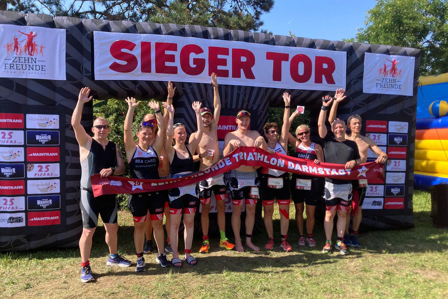 Gruppe von Triathleten feiert unter einem Siegerbogen mit einem Banner für den "Darmstadt Triathlon".