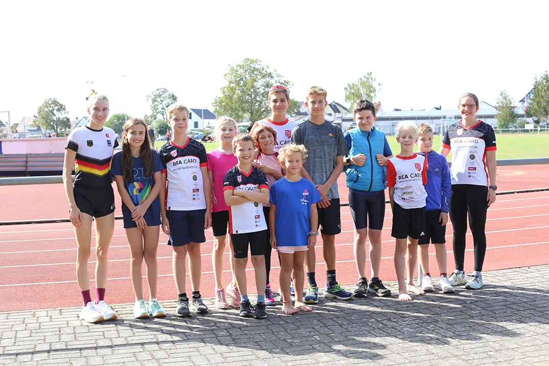 Gruppe von zehn Kindern und Jugendlichen in Sportkleidung steht auf einer Laufbahn im Freien.