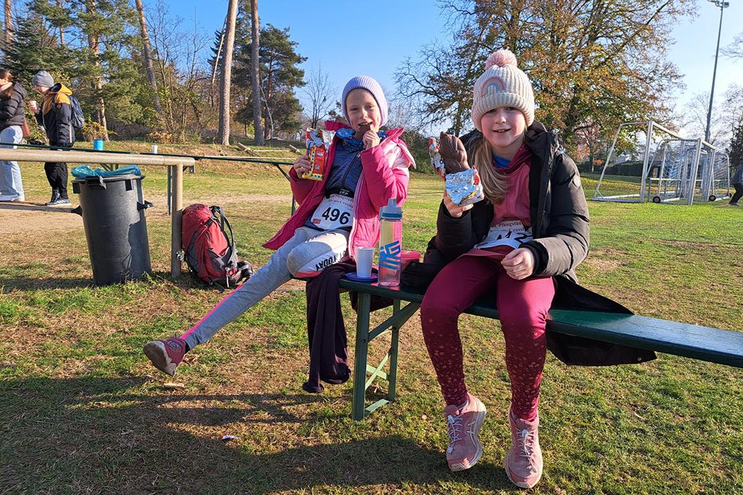 Zwei Mädchen sitzen auf einer Bank, essen Snacks und genießen den Sonnenschein auf einem Sportplatz.