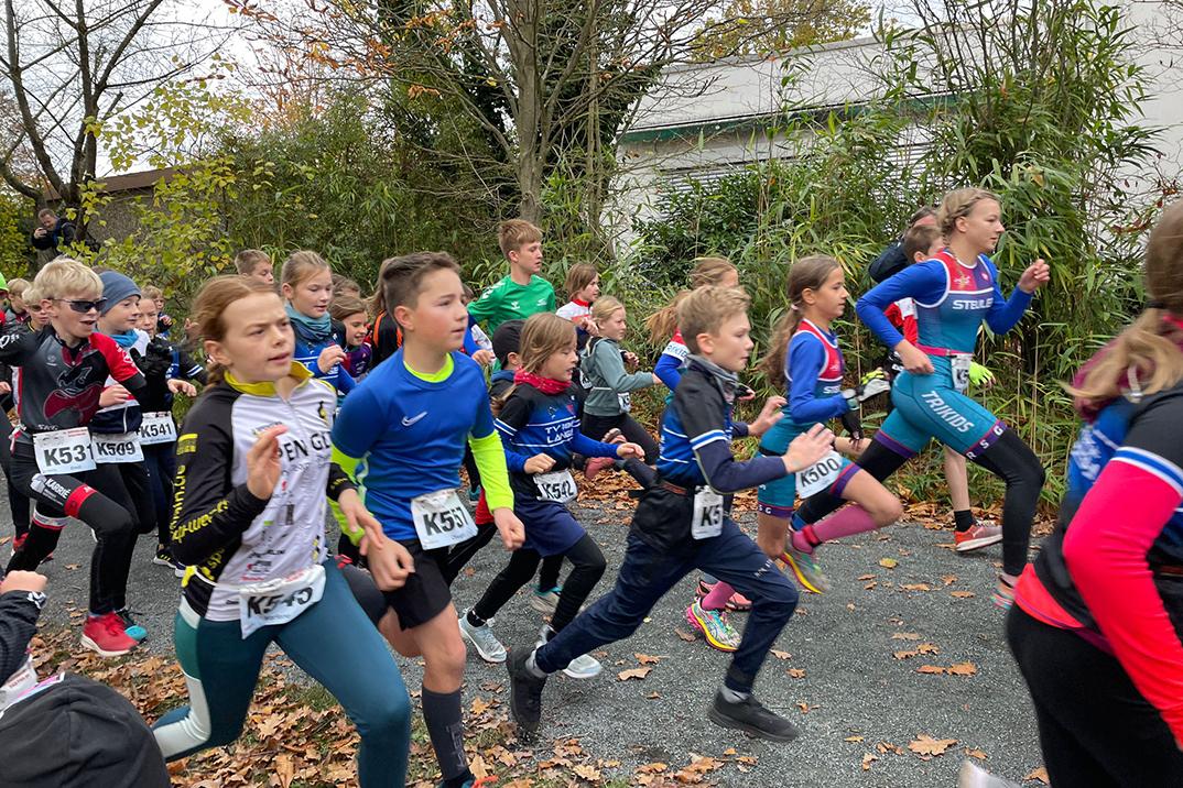 Gruppe von Kindern in Sportkleidung beim Laufen auf einem Weg, umgeben von Bäumen und herbstlichem Laub.