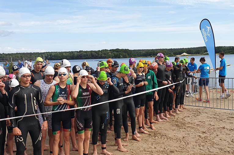 Wettkampfteilnehmer in Neoprenanzügen und Schwimmkappen warten an einem Strand vor einer Wasserfläche auf den Start.
