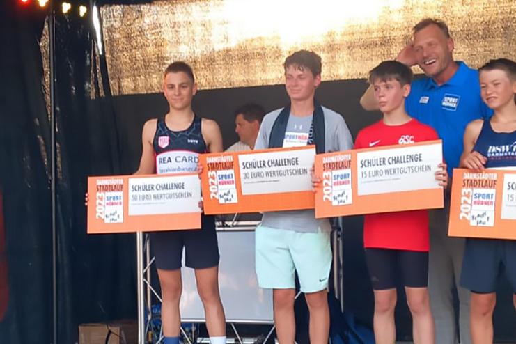 Vier Gewinner der Sch&uuml;ler-Challenge halten ihre Gutscheine auf einer B&uuml;hne, w&auml;hrend ein Moderator anspricht.