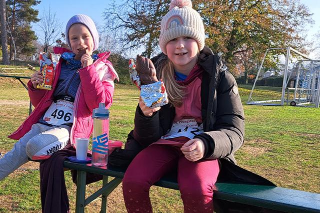 Zwei Mädchen sitzen auf einer Bank, essen Snacks und genießen den Sonnenschein auf einem Sportplatz.