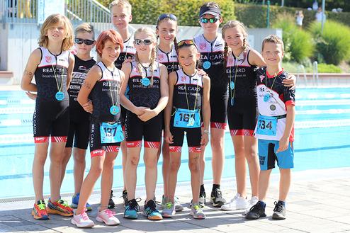 Gruppe von zehn Kindern in Sportanzügen, lächelnd, vor einem Schwimmbecken, einige mit Medaillen um den Hals.